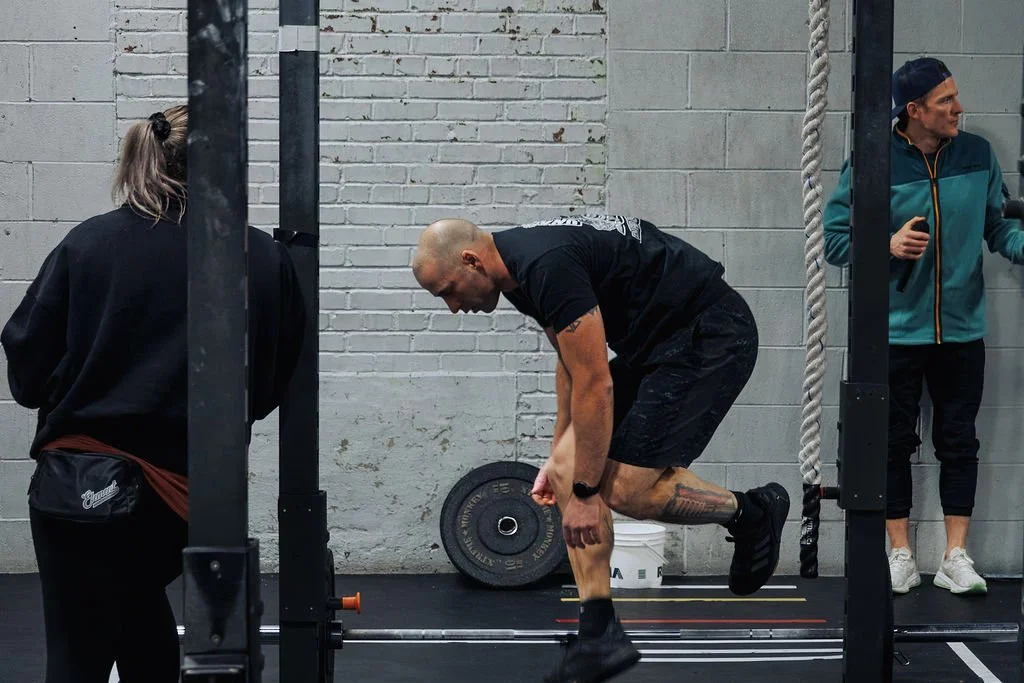 Man performing a box jump exercise in a gym, with two women observing nearby.