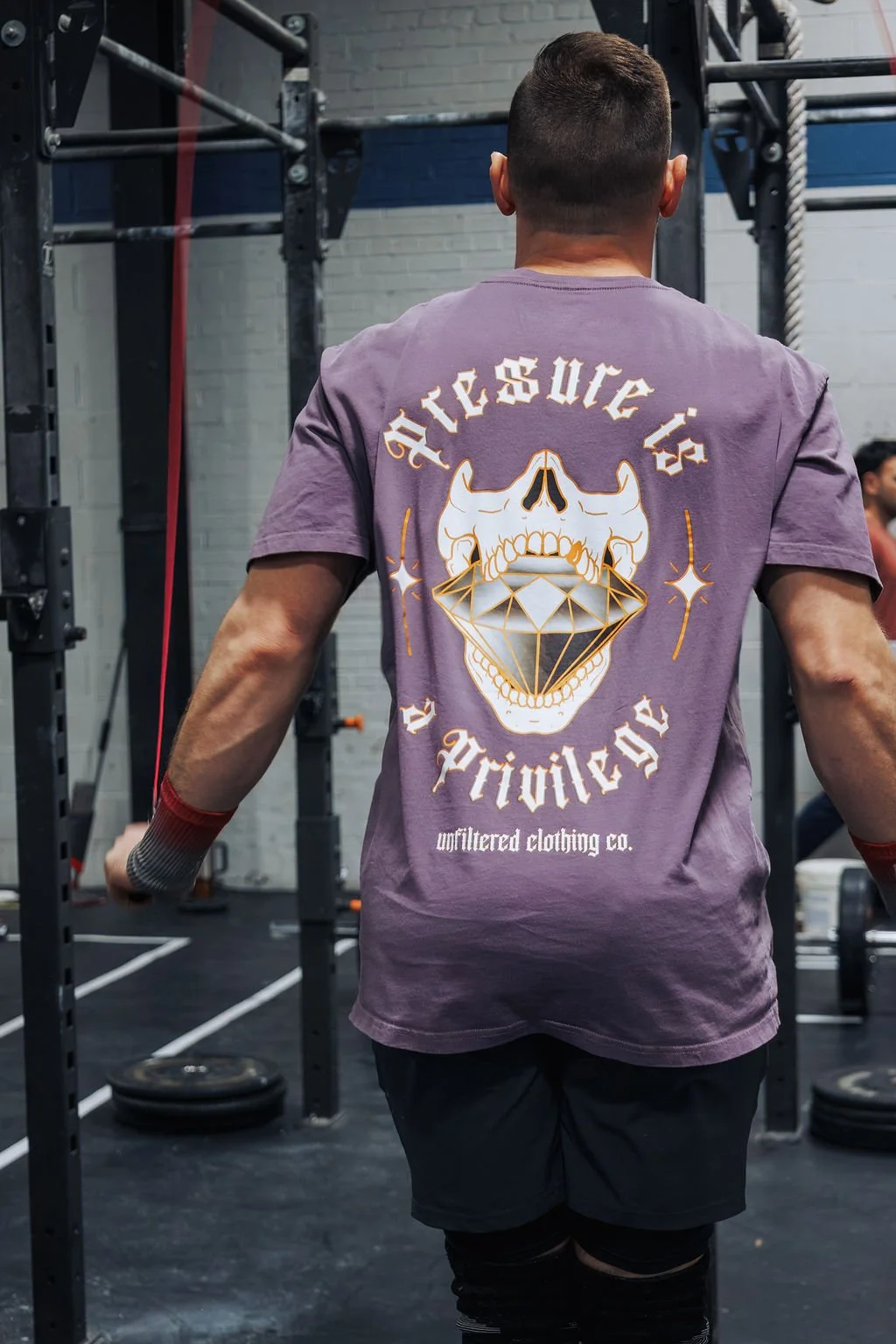 Back of a person in a purple t-shirt with a skull and diamond graphic and the text "Acceptance is Privilege" at a gym.