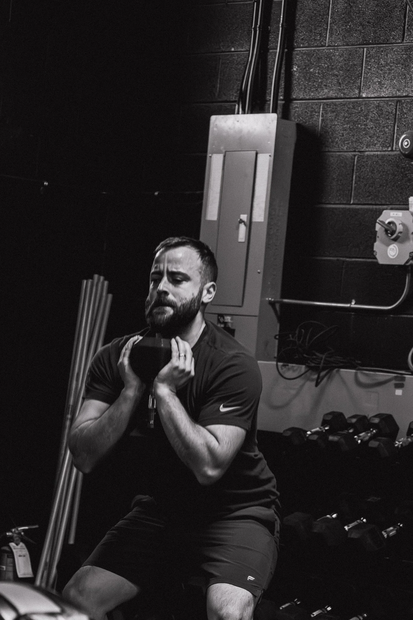 Man in workout attire performing a seated exercise with a weight plate on his shoulders in a gym setting, black and white photo.