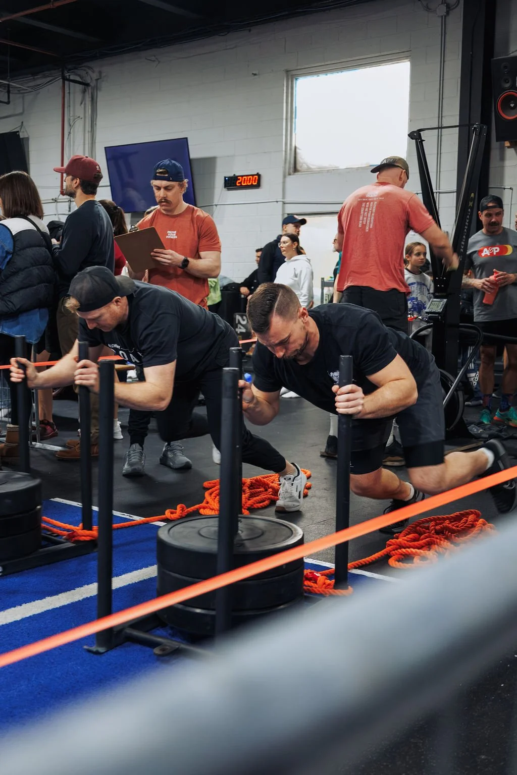 Two men participating in a workout at a gym, doing push-ups with weighted sleds on their backs, surrounded by a group of people and gym equipment.