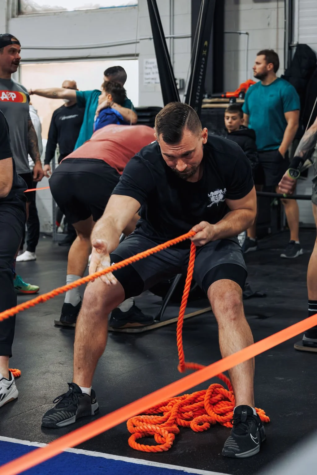 A man participating in a strongman workout, pulling a thick orange rope with a determined expression in a gym filled with other people and gym equipment.