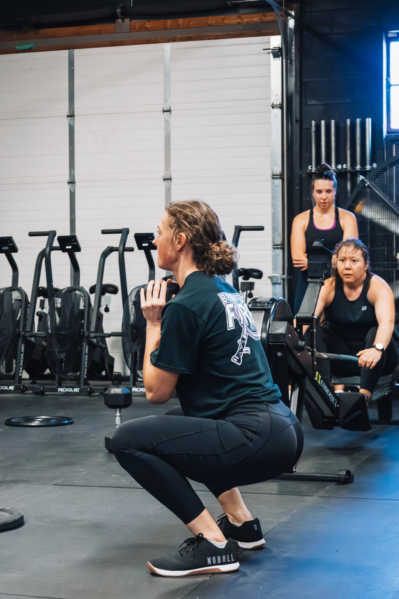 Woman in black workout clothes squatting during a fitness class in a gym with two women in the background observing.
