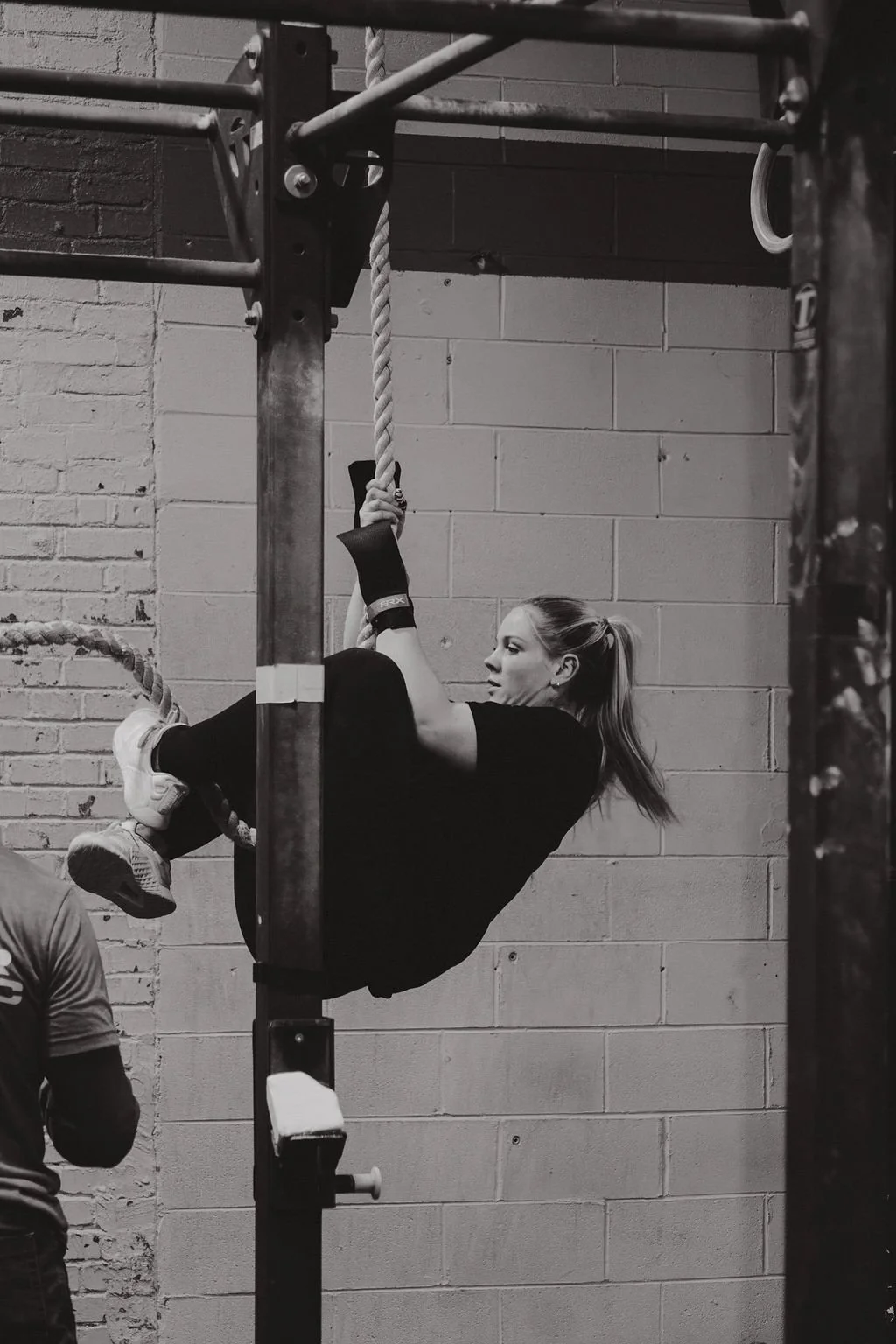 A woman performing a hanging exercise on gym ropes, holding one rope with her right hand, in a gym with brick and concrete walls.