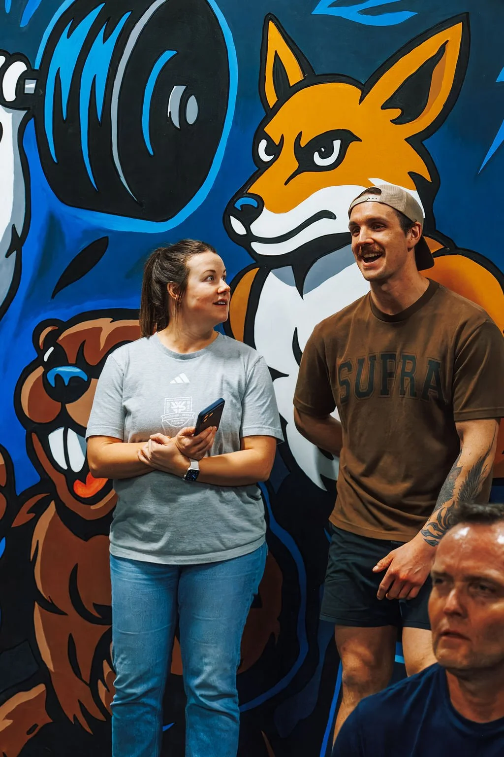 Two people talking in front of a colorful mural of a fox, bear, and wolf with a large speaker.