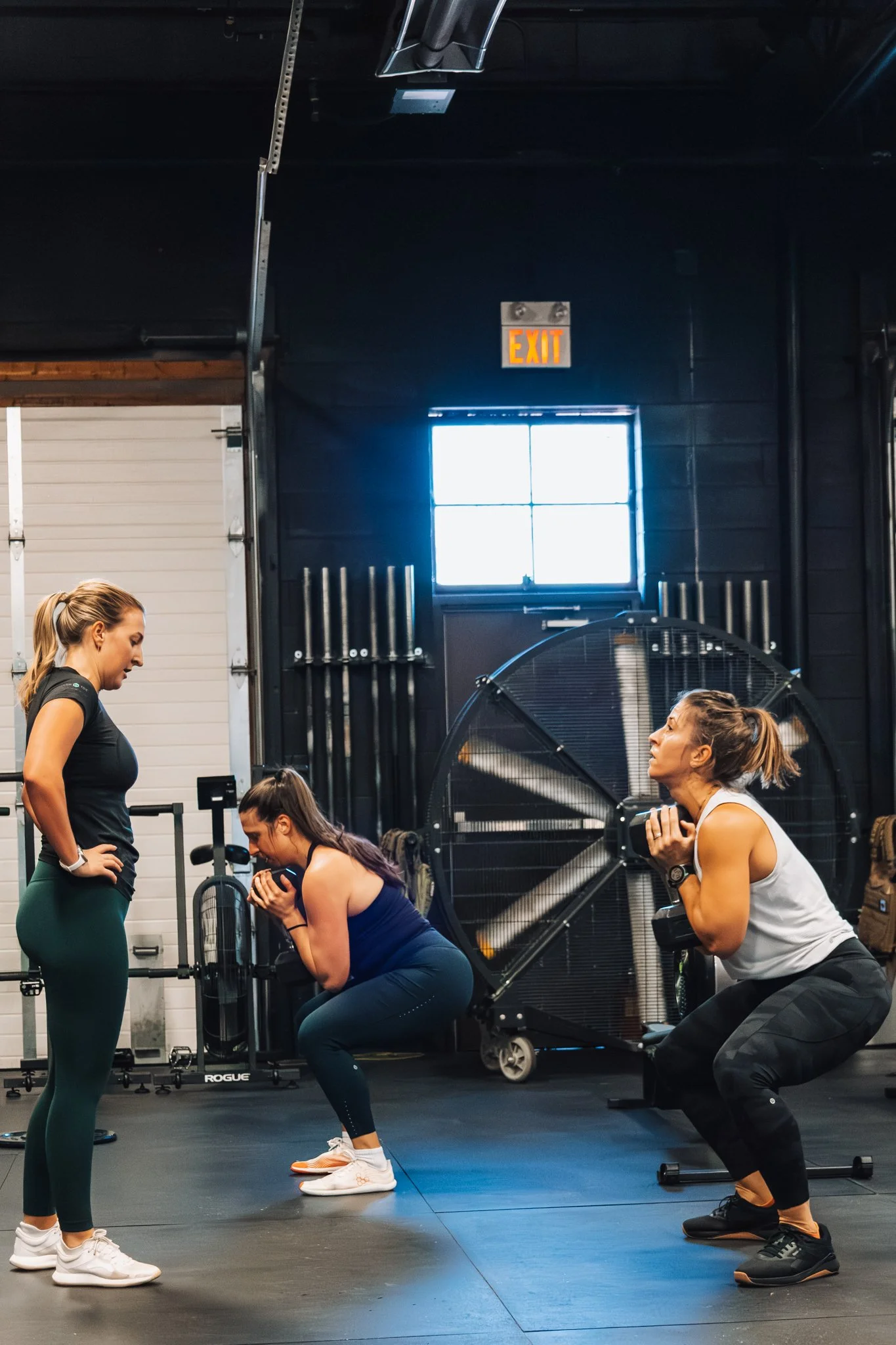 Three women exercising in a gym. One woman is standing and coaching, the other two women are squatting with weights and resistance bands.