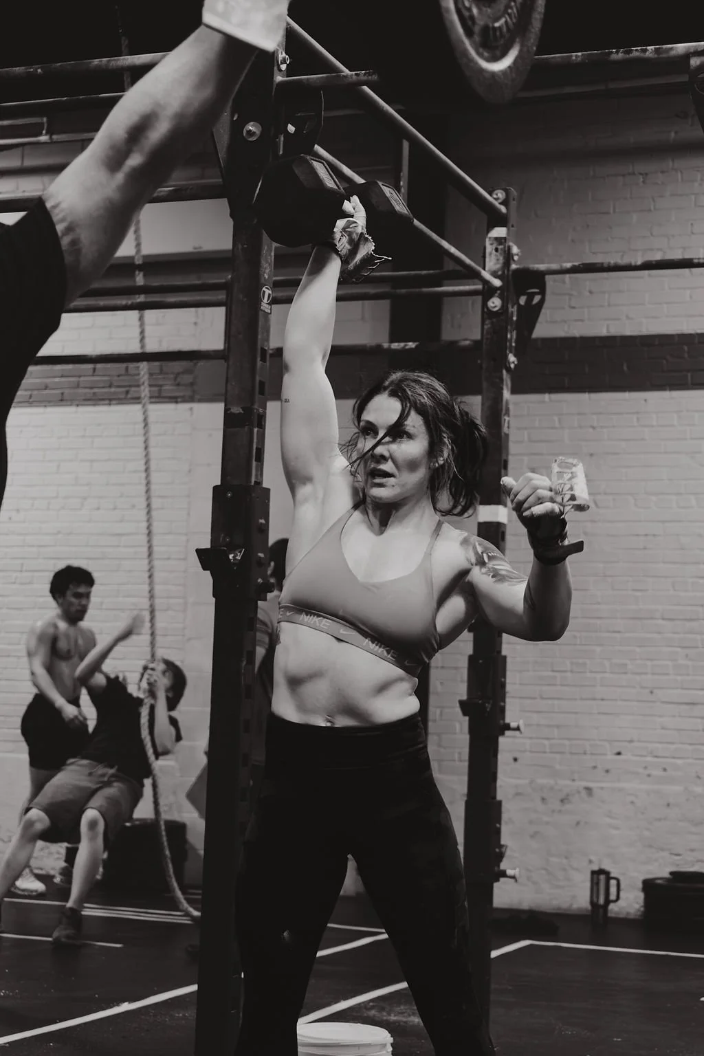 A woman in workout clothes lifting a dumbbell overhead in a gym, with children playing and running in the background.