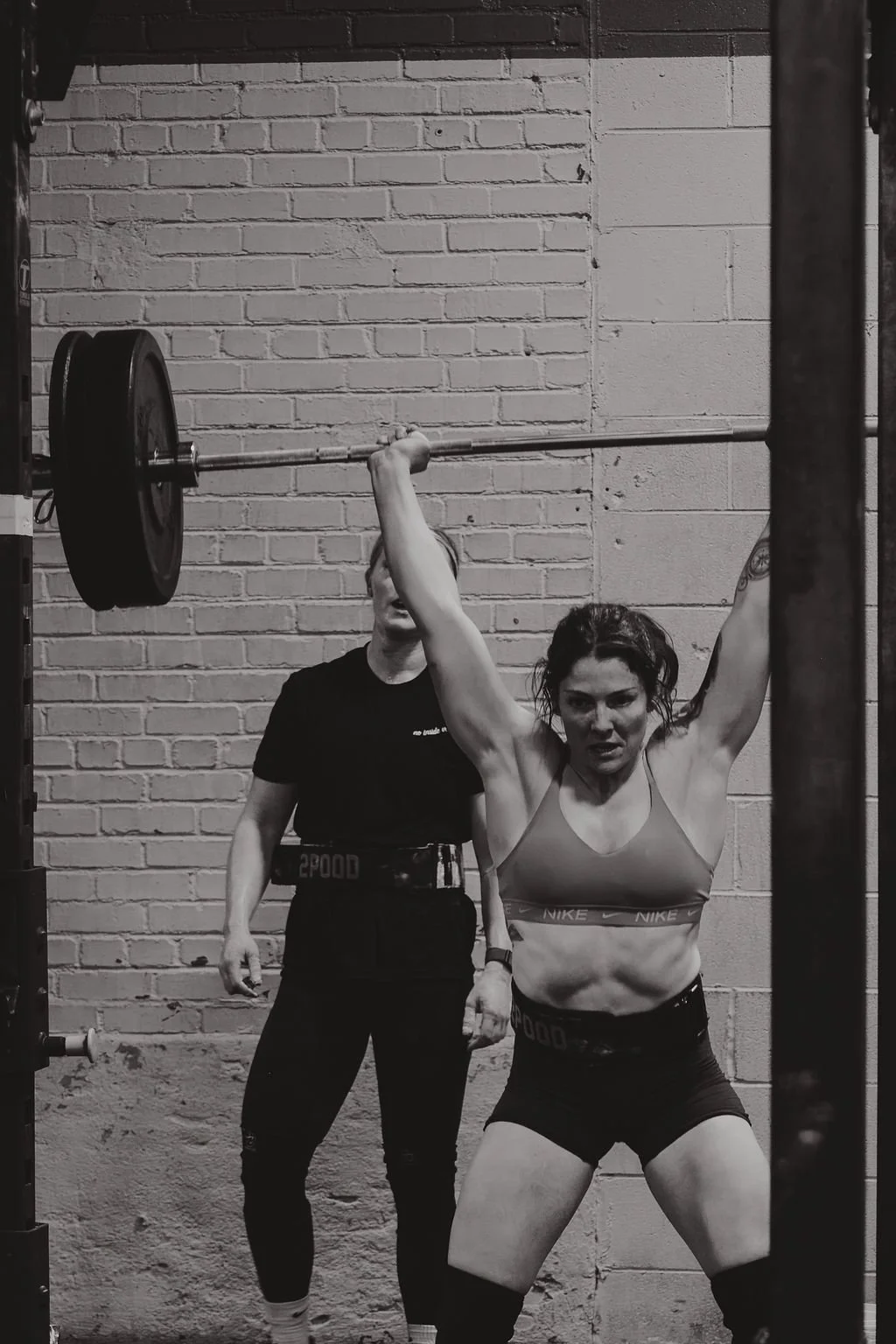A woman with tattoos wearing sportswear performs a lifting exercise with a barbell in a gym, overseen by a coach or trainer standing behind her against a brick wall.