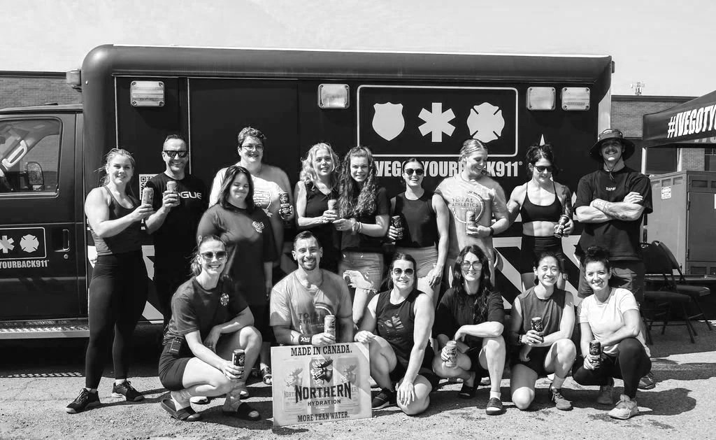 Group of people posing outdoors near a black emergency vehicle, holding drinks, some wearing athletic clothing.