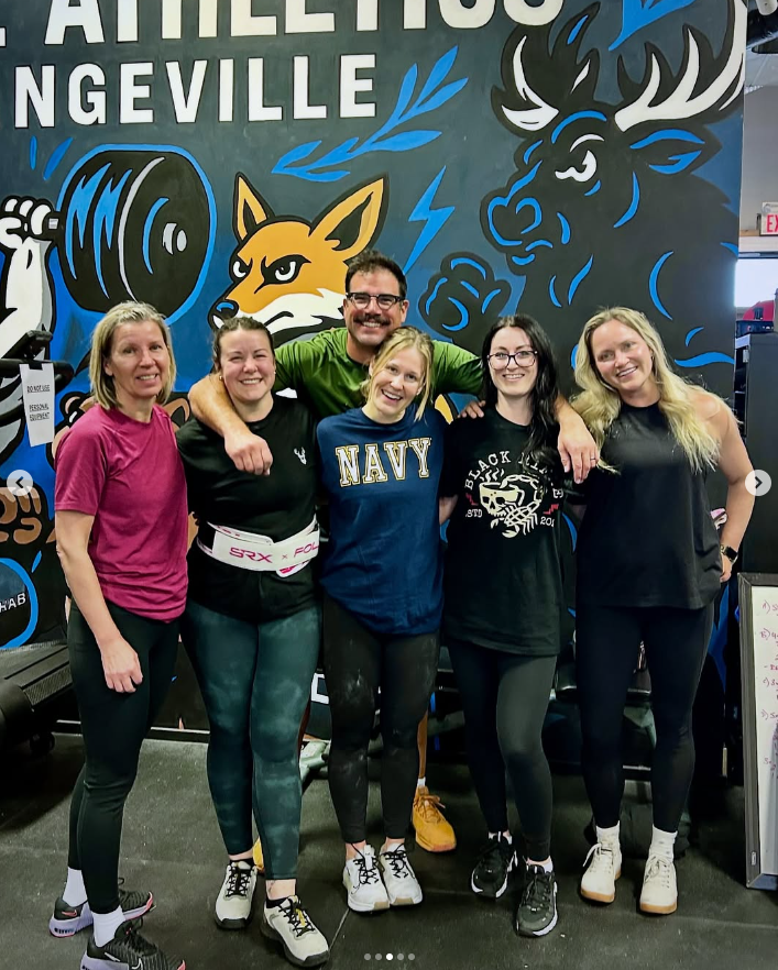 Group of five women and one man smiling and posing together in a gym, with a colorful mural of a fox, a moose, and weights in the background.