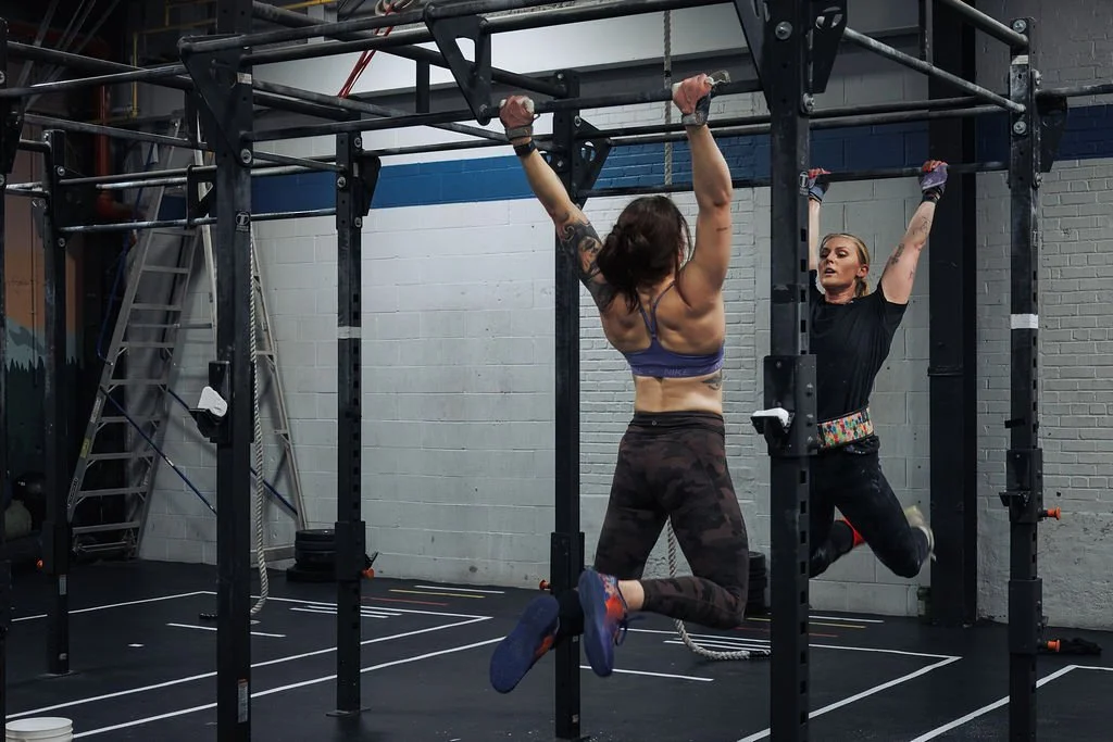 Two women working out on a pull-up and hanging leg raise station in a gym. One woman is hanging from the pull-up bar, and the other is doing a hanging leg raise.