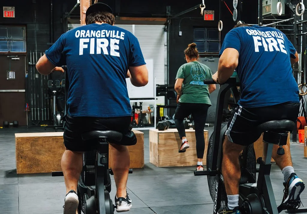 Three people working out in a gym, two on exercise bikes wearing blue shirts with 'Orangeville Fire' on the back, and one on a wooden box doing step exercises, in a gym with fitness equipment and squat racks.
