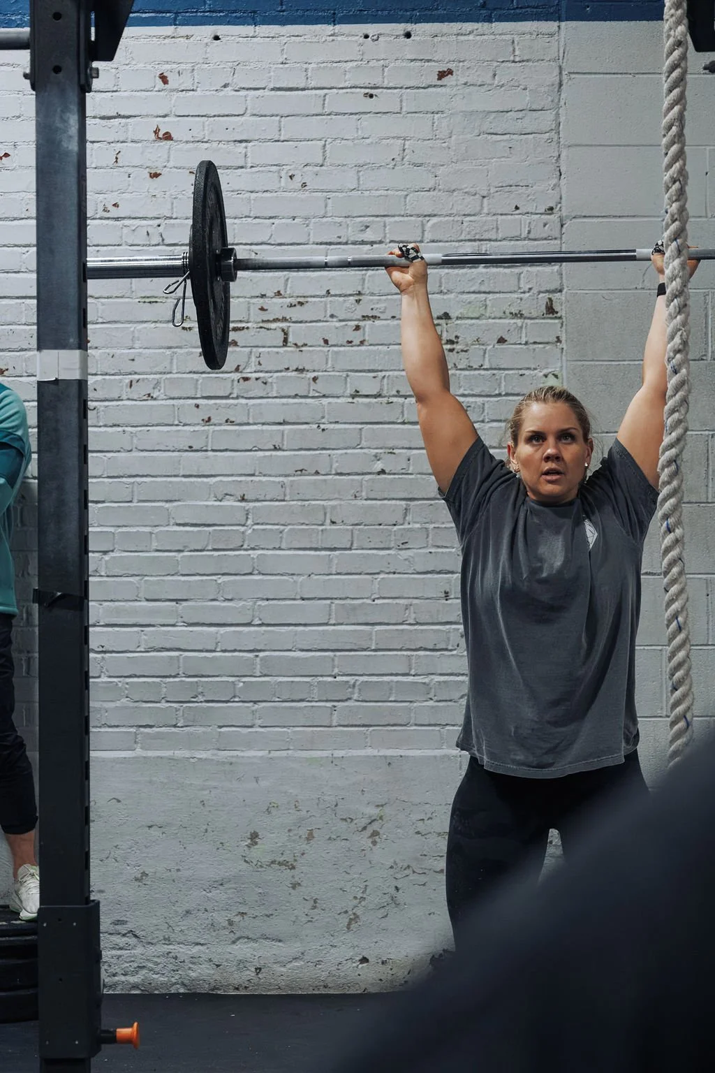 Woman lifting a barbell overhead during a workout at a gym with a white brick wall background.