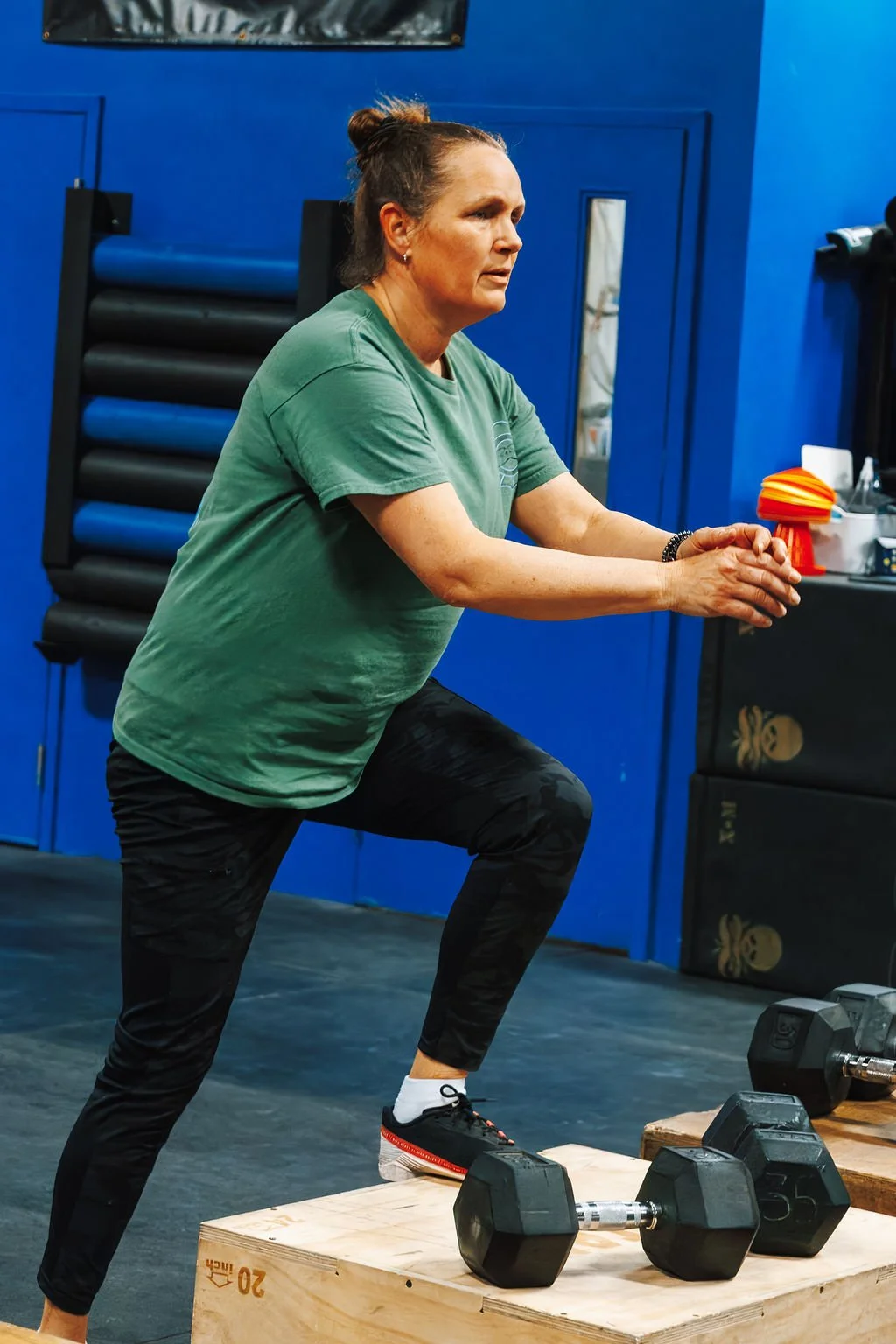 A woman in exercise attire performing a lunge exercise with dumbbells placed on a wooden platform in a gym with blue walls.