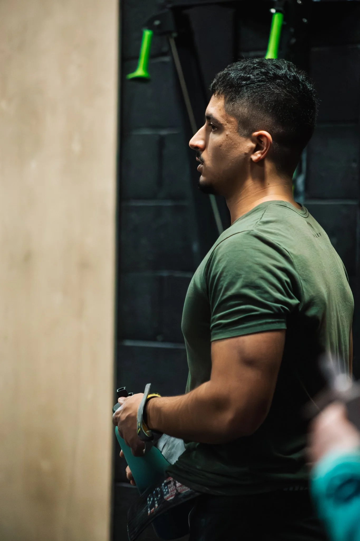 A man standing indoors wearing an olive green t-shirt, holding a water bottle, with black brick wall and gym equipment in the background.