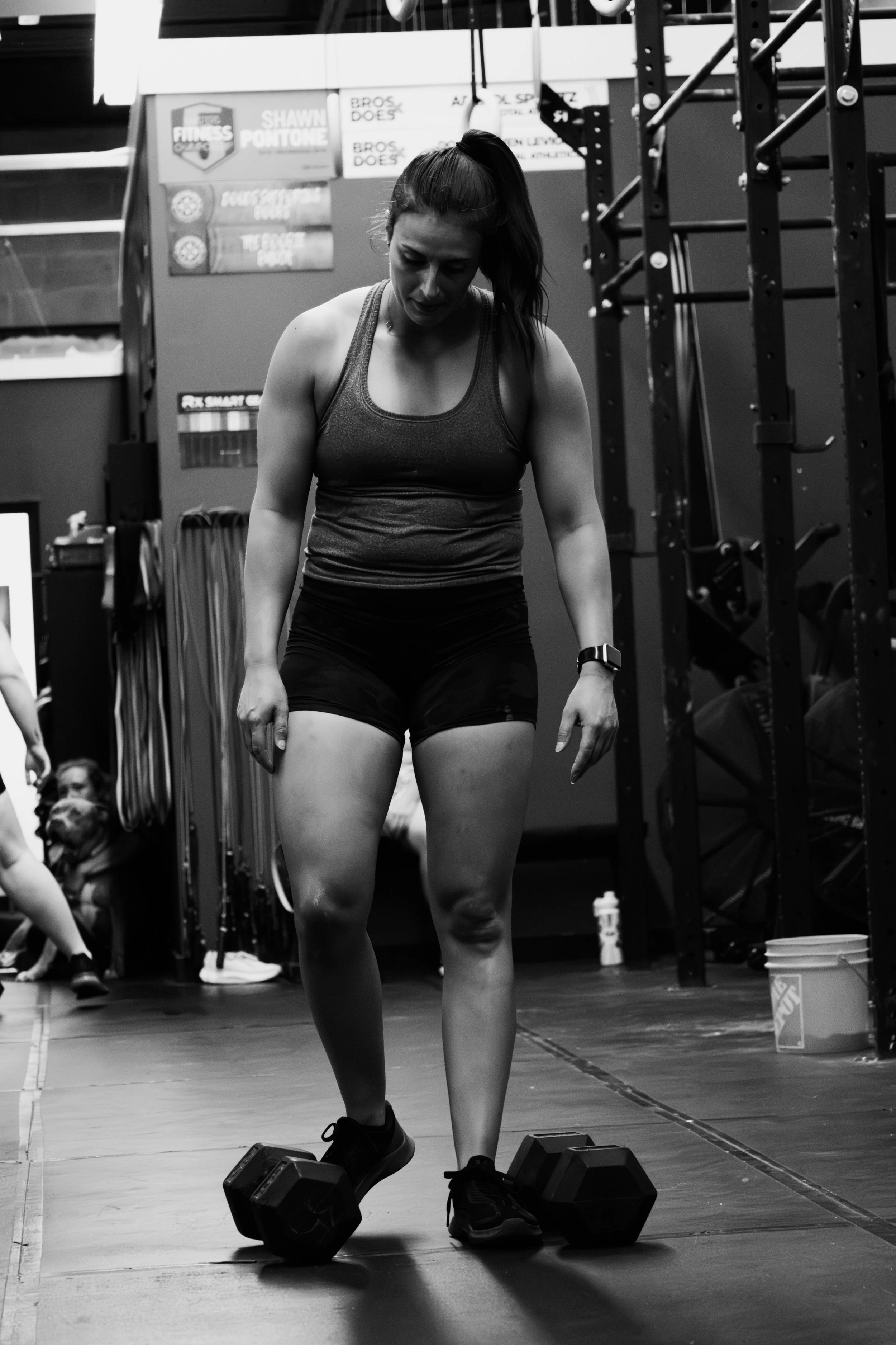 A woman in athletic wear is standing in a gym with a kettlebell on each side of her feet, looking downward at her feet.