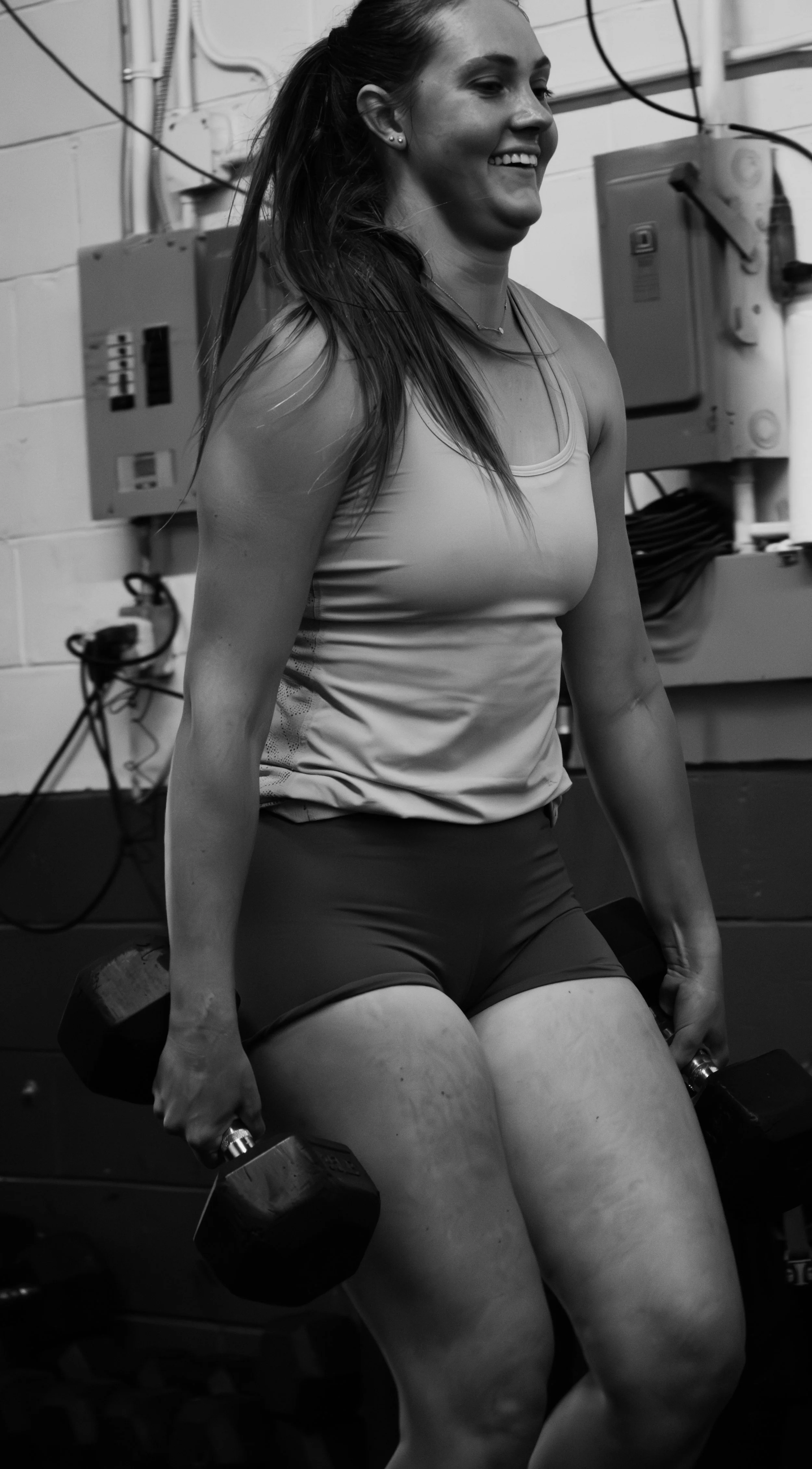 A woman in workout attire smiling while holding dumbbells in a gym setting.