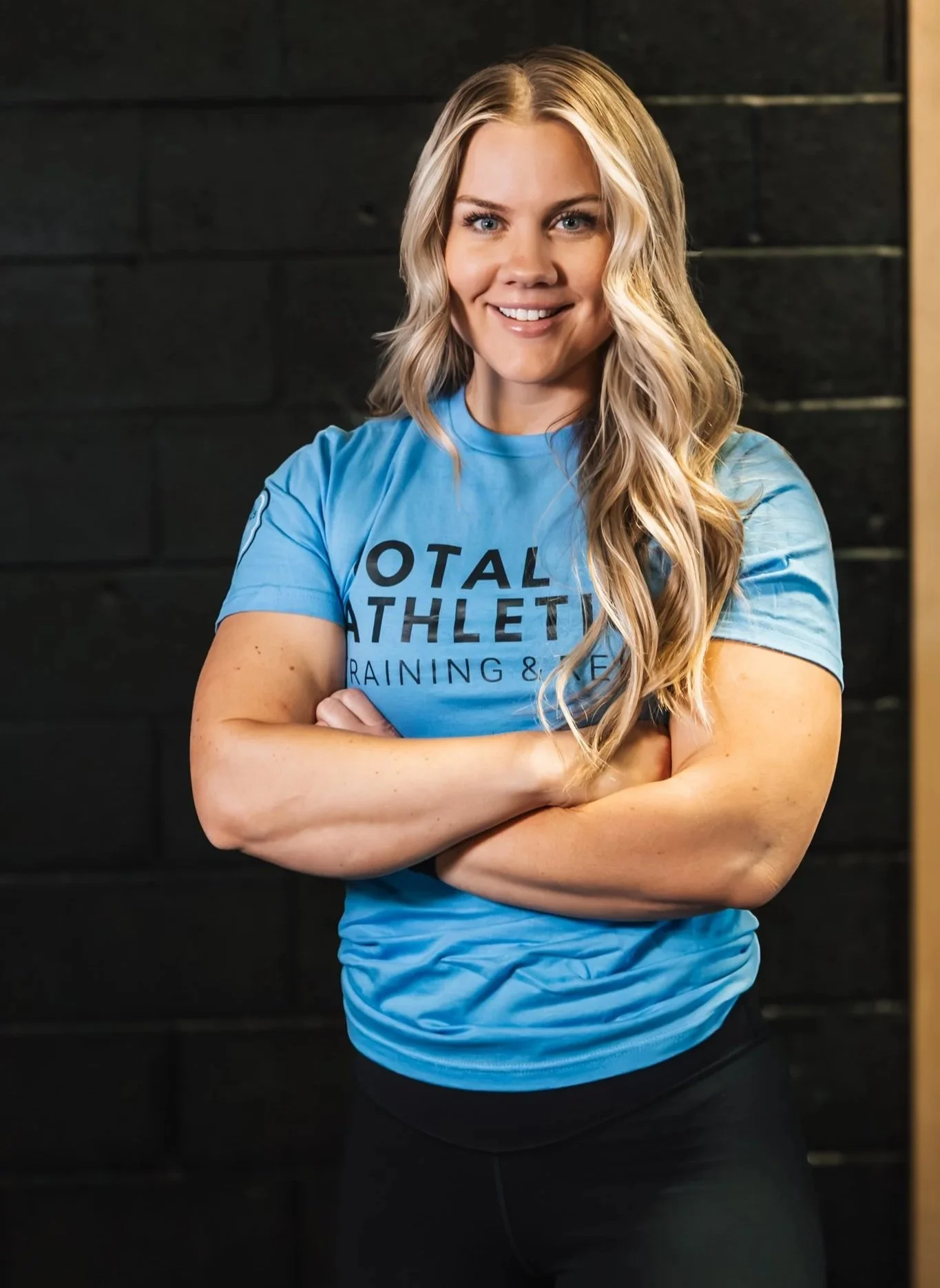 A young woman with blonde wavy hair, wearing a black t-shirt and light blue jeans, standing with crossed arms in front of a wall with gym equipment. She is smiling.