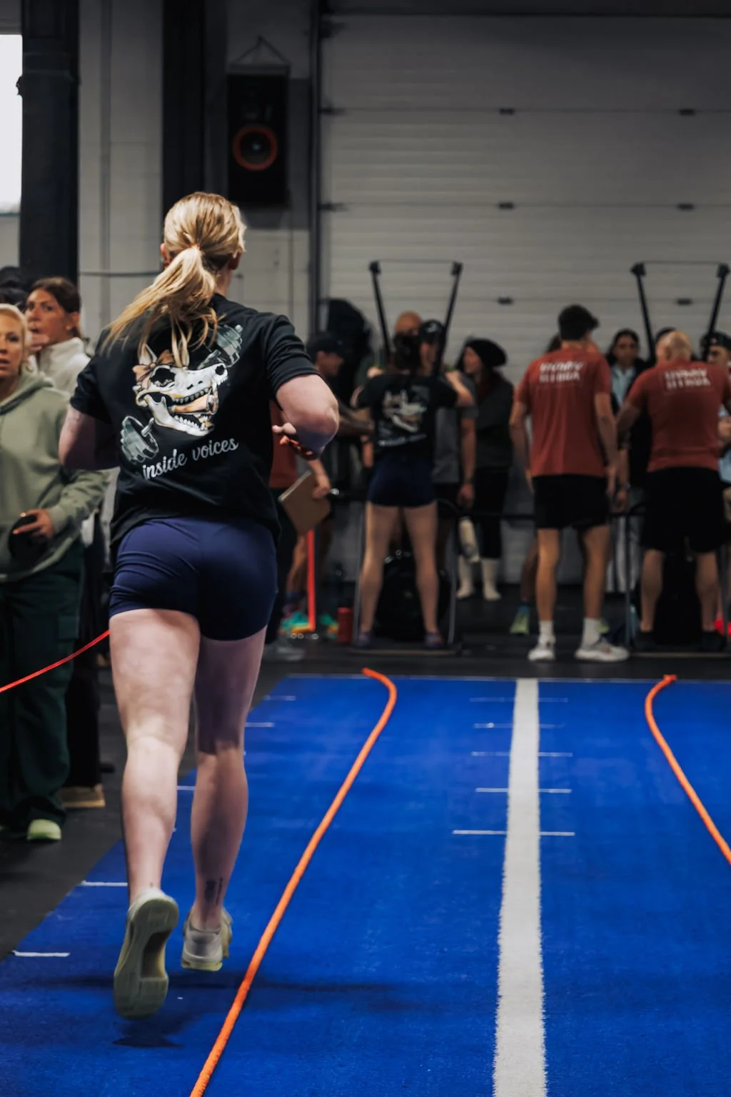 A woman running on a blue track in an indoor gym, with a crowd of people in the background, some wearing event shirts, and a large speaker mounted on the wall.
