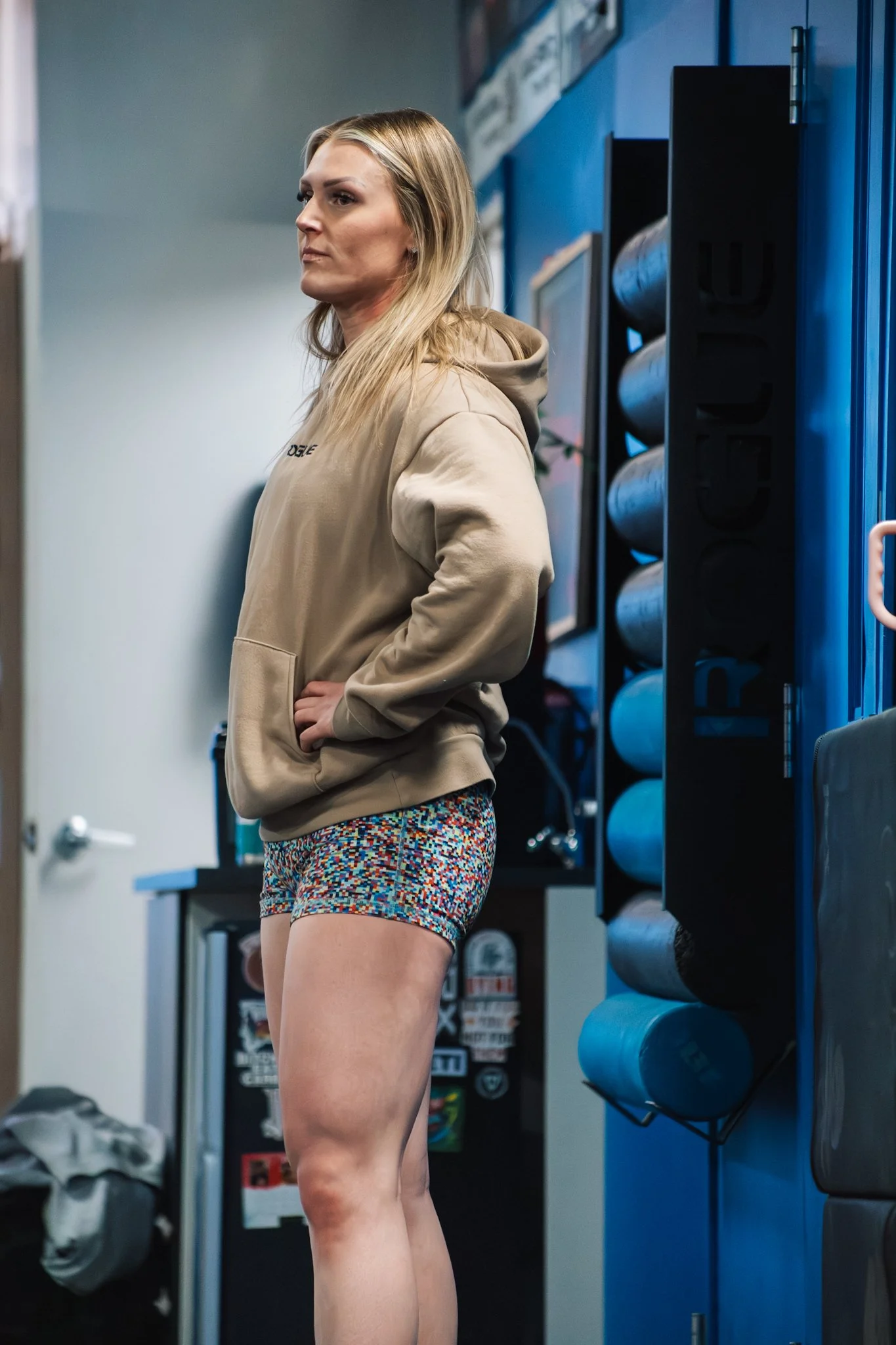A woman with long blonde hair wearing a beige hoodie and colorful shorts standing in a gym.