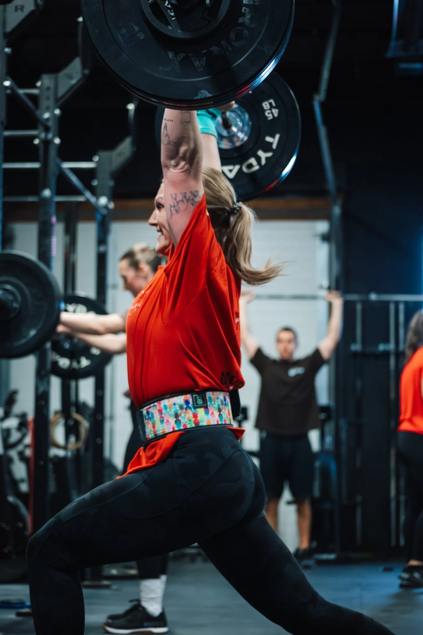 A woman lifting a heavy barbell overhead during a weightlifting workout in a gym, with other people exercising in the background.