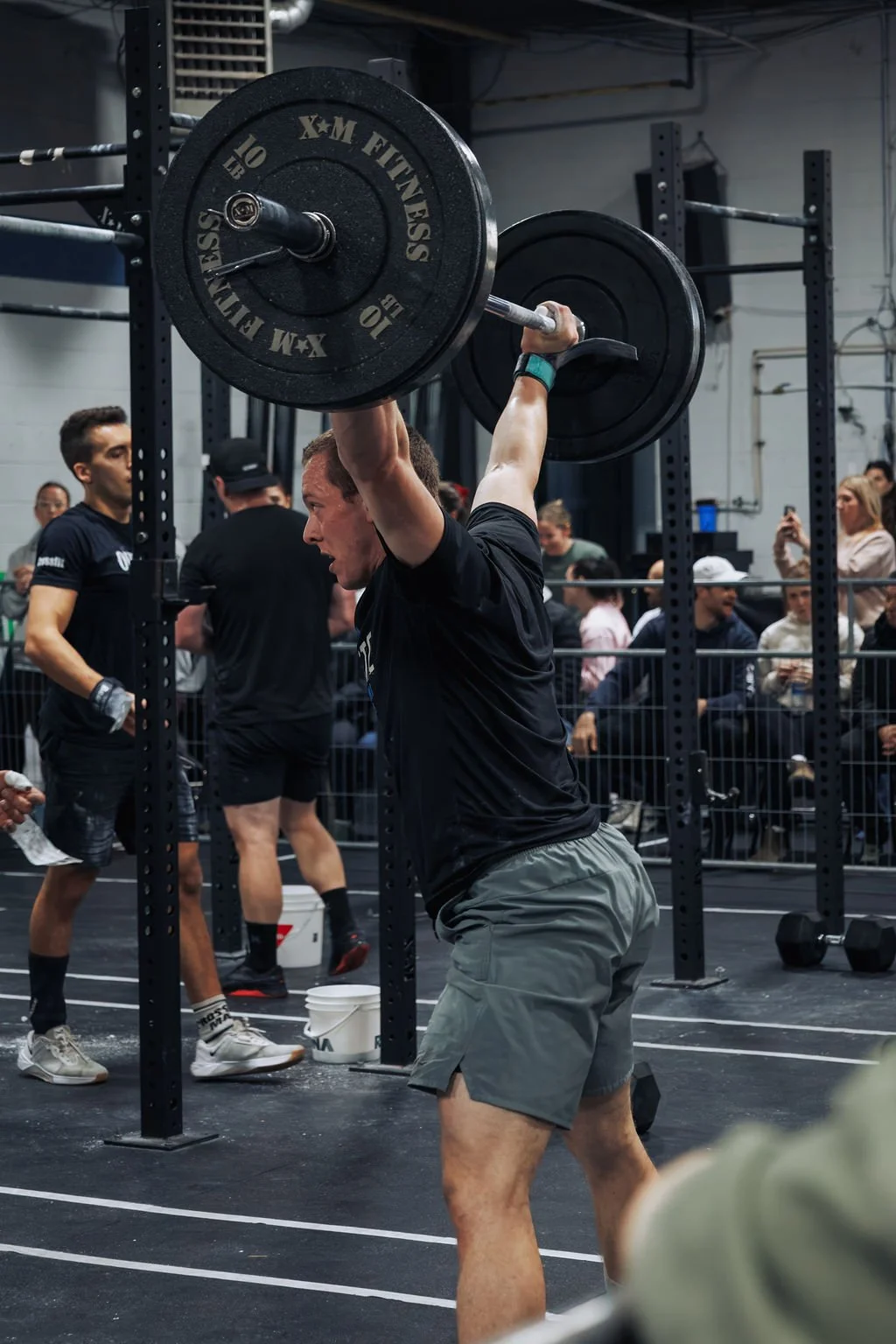 A man lifting a large barbell above his head during a weightlifting competition in a gym, with spectators and other athletes in the background.