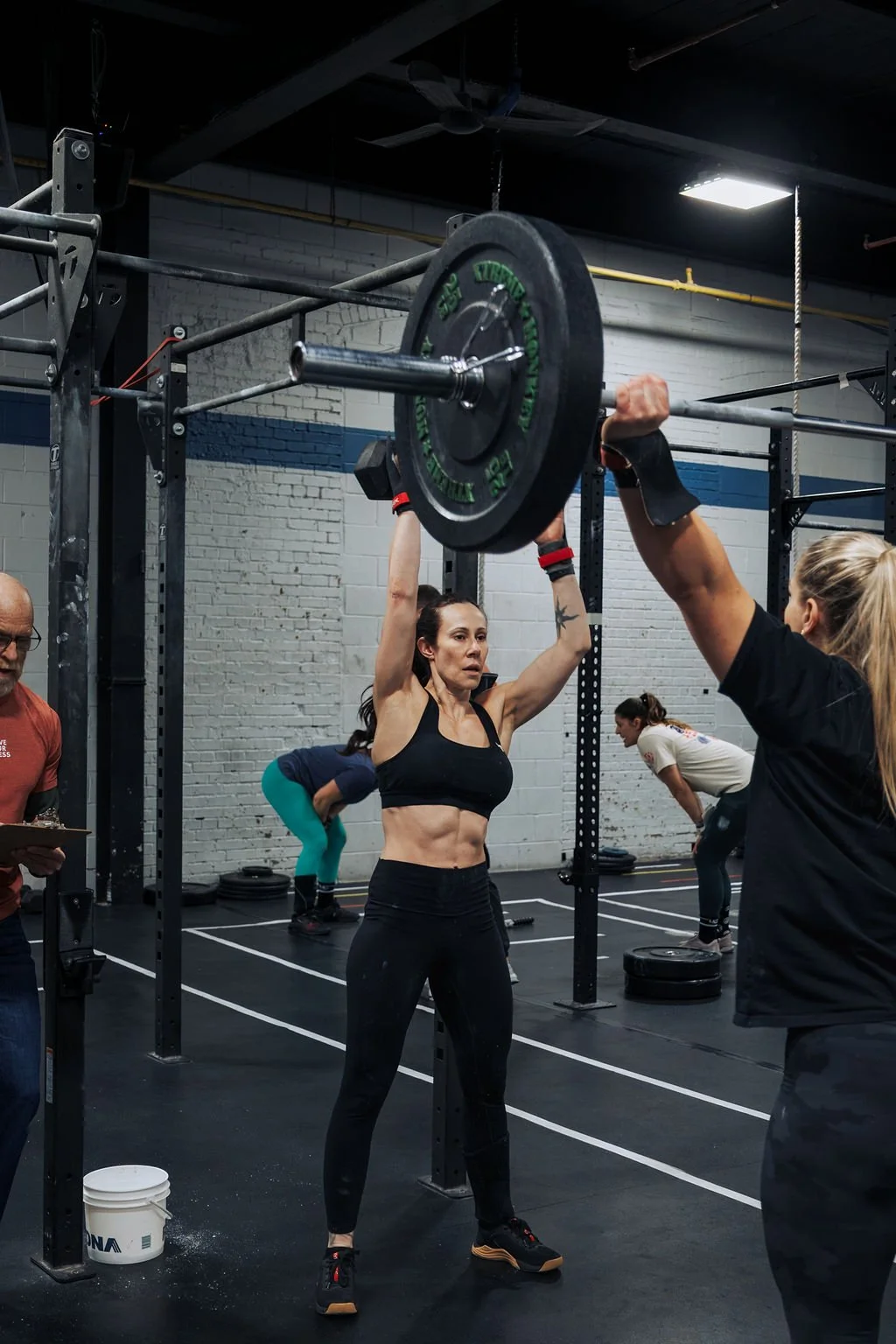 A woman lifting a barbell with weights during a workout at a gym, with other people exercising in the background.