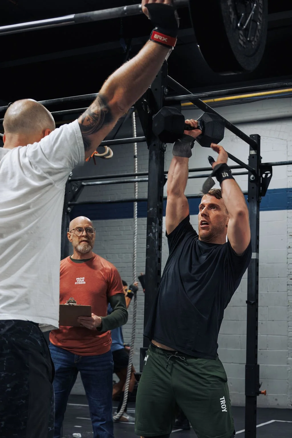 A man in a black shirt doing a pull-up at a gym, with two other men observing and one holding a clipboard.