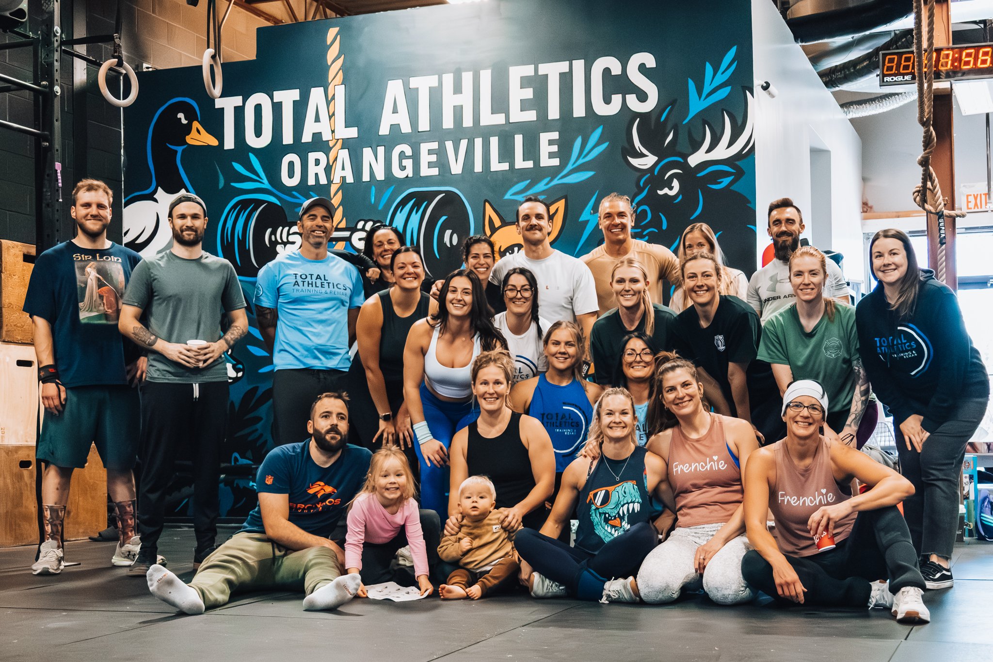 Group of people at the gym in front of a wall with 'Total Athletics Orangeville' logo, diverse ages, smiling, some children, gym equipment visible.