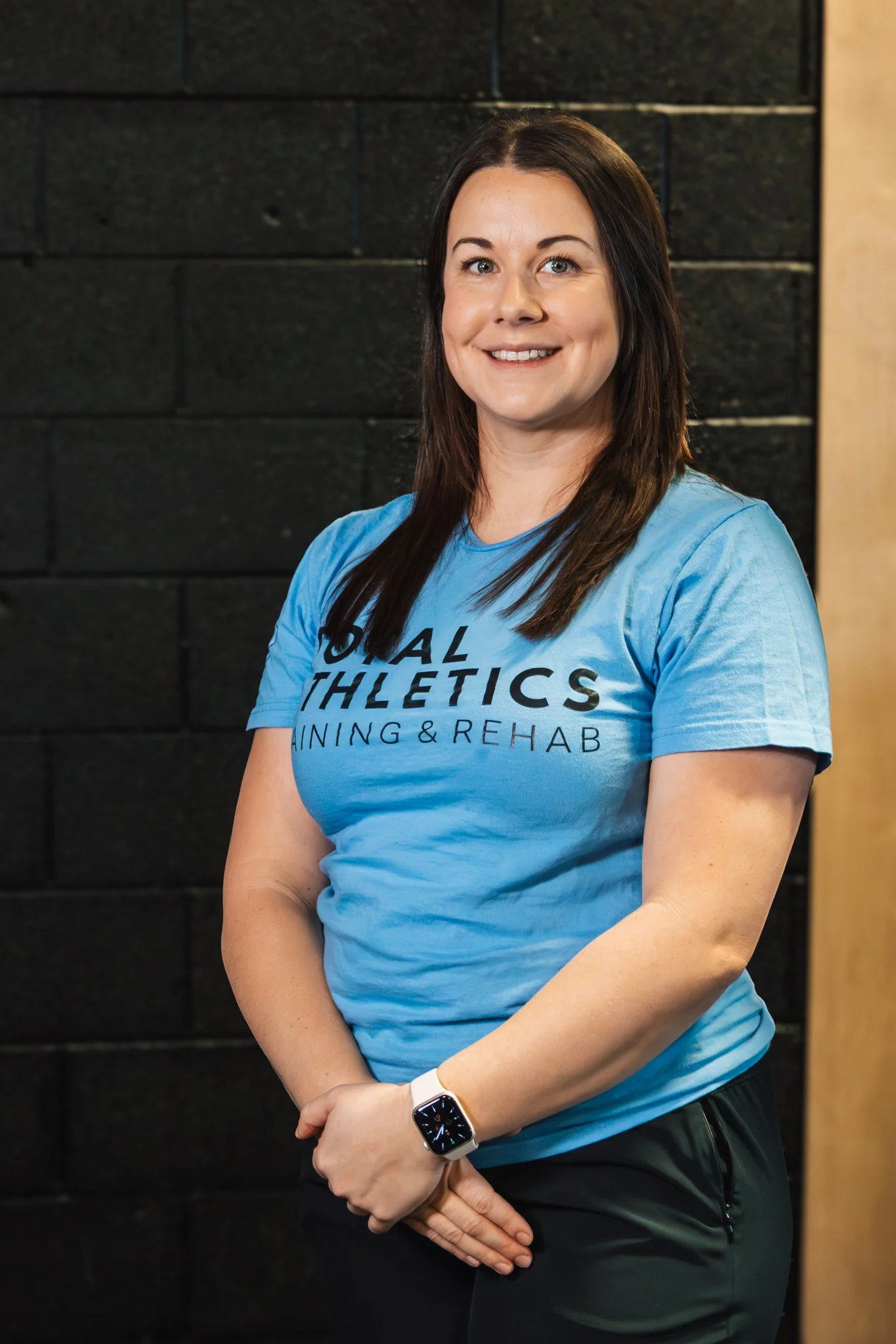 A woman with shoulder-length brown hair, wearing a black t-shirt and blue jeans, standing with arms crossed in front of a white brick wall and a black fitness fan.