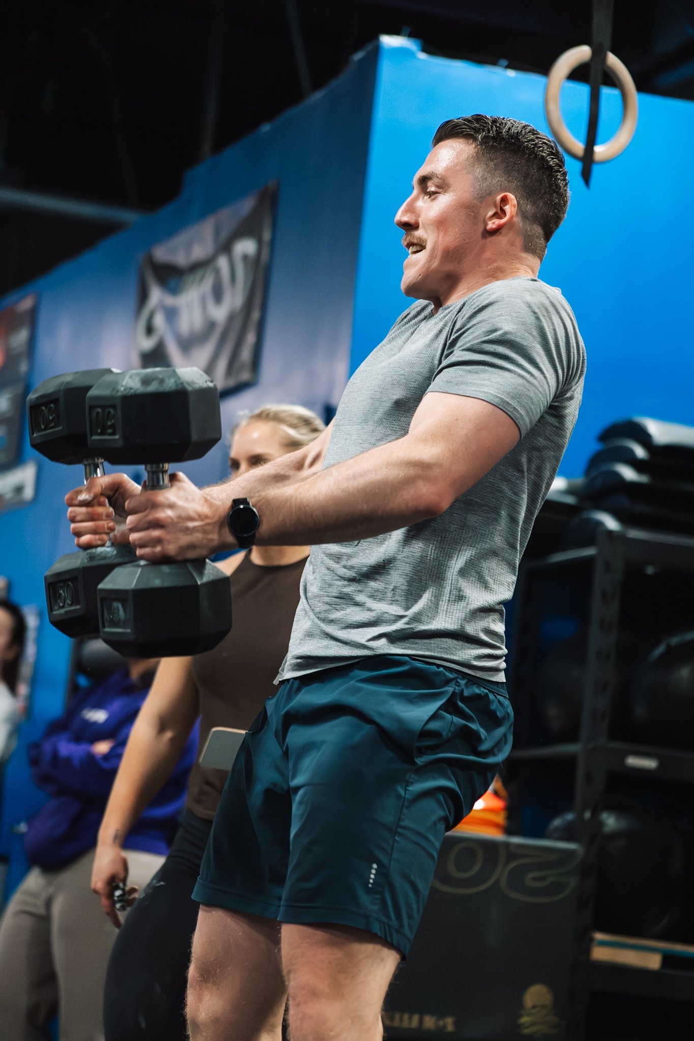A man lifting a dumbbell during a workout in a gym, with a woman and other people in the background.