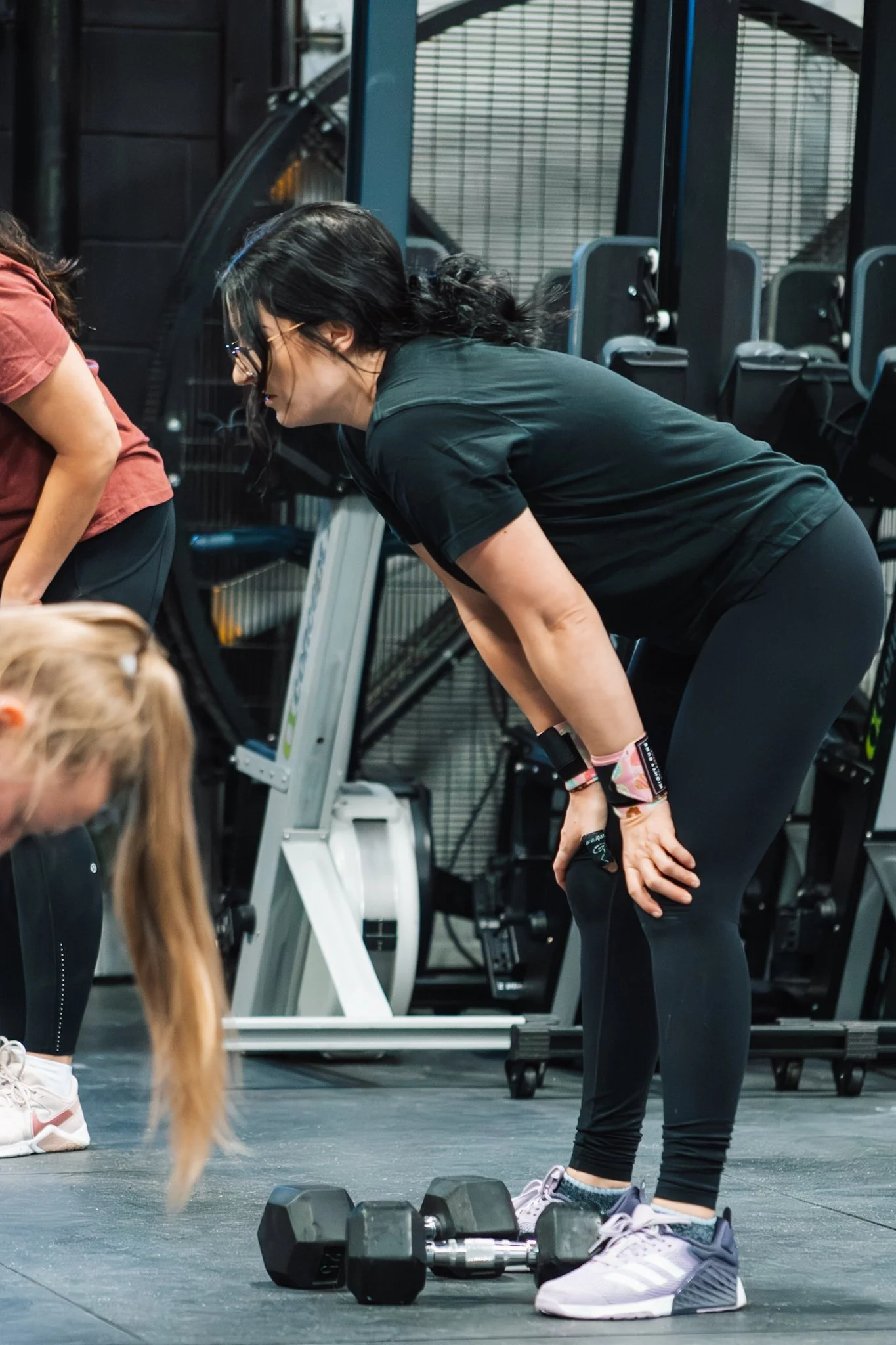 A woman with black hair, glasses, and wearing black workout clothes, bending over during a workout session in a gym.