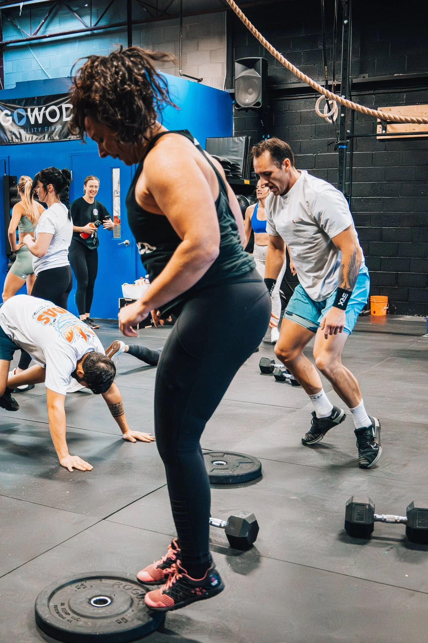 Group of people working out in a gym, some doing push-ups on the floor, others jumping or standing near weight plates and dumbbells.