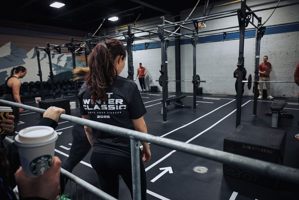 People in a gym preparing for a workout, with racks and weights, and a woman in the foreground wearing a black shirt with white text.