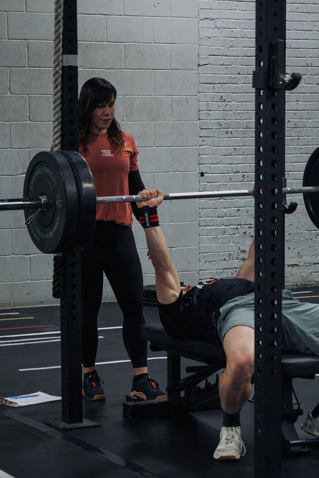 A woman spotting a man performing a bench press in a gym with industrial brick walls.