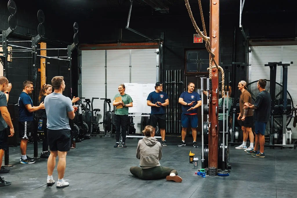 Group of people gathered in a gym, listening to a woman holding a clipboard, with gym equipment and mats around them.