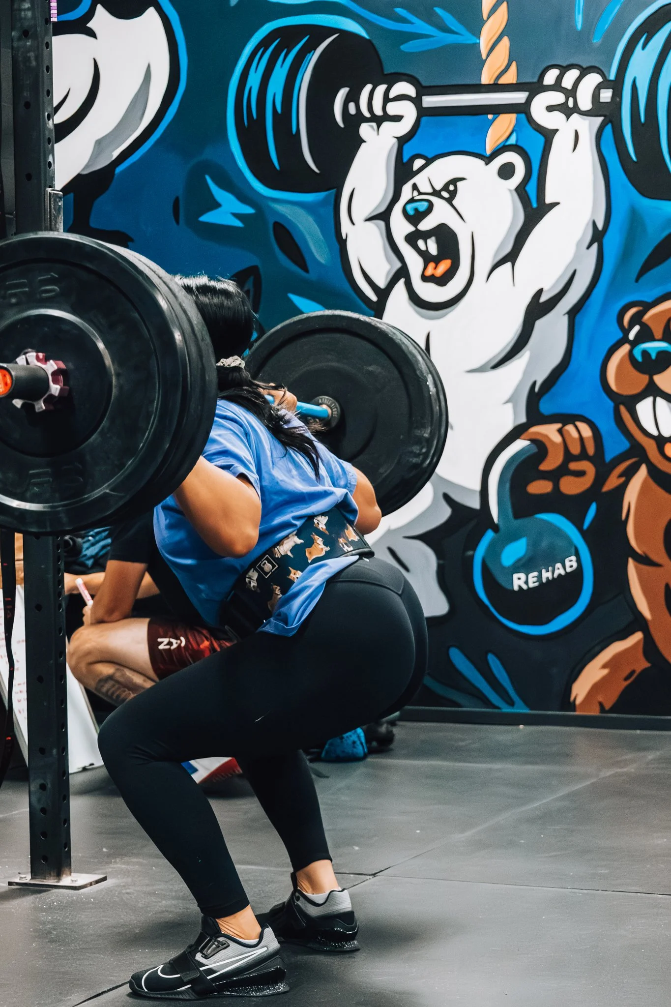 A woman lifting a barbell in a gym with colorful animal murals on the wall, including a bear and a tanuki, and a kettlebell with the word 'REHAB' written on it.