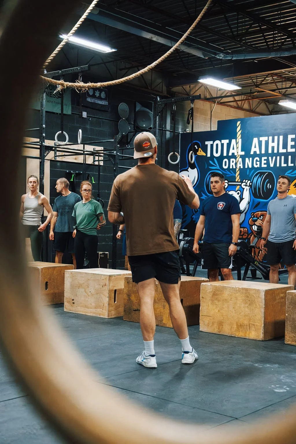A fitness class taking place in a gym with people listening to an instructor. The gym has a blue sign that reads 'Total Athlete Orangeville' and health and safety signs on the wall.