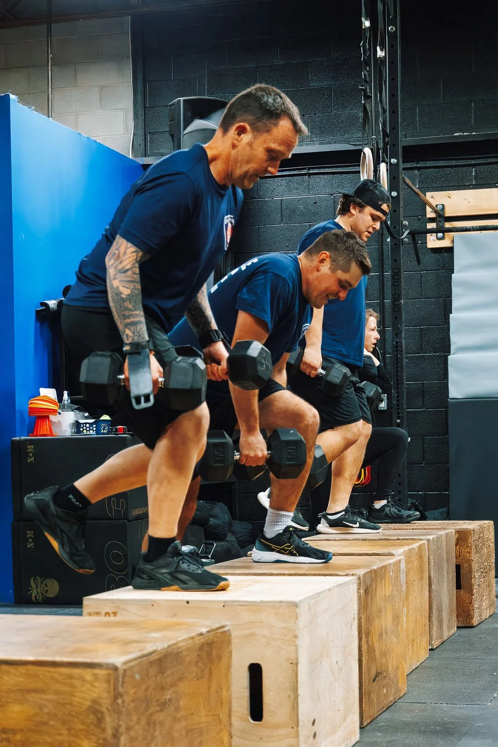 Four men perform wall squats with dumbbells in a gym. They are in a row, holding dumbbells, with wooden blocks under their feet, black wall, and gym equipment in the background.