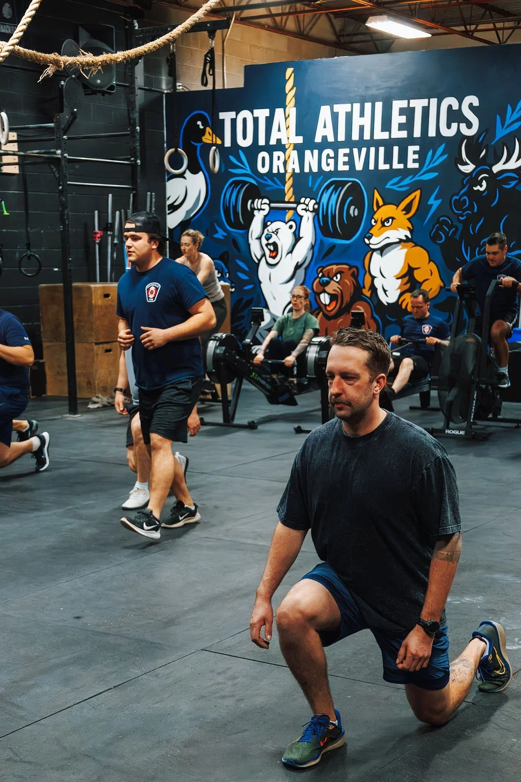 People working out in a gym inside Total Athletics Orangeville, with a large wall mural featuring cartoon animals and the gym's name.