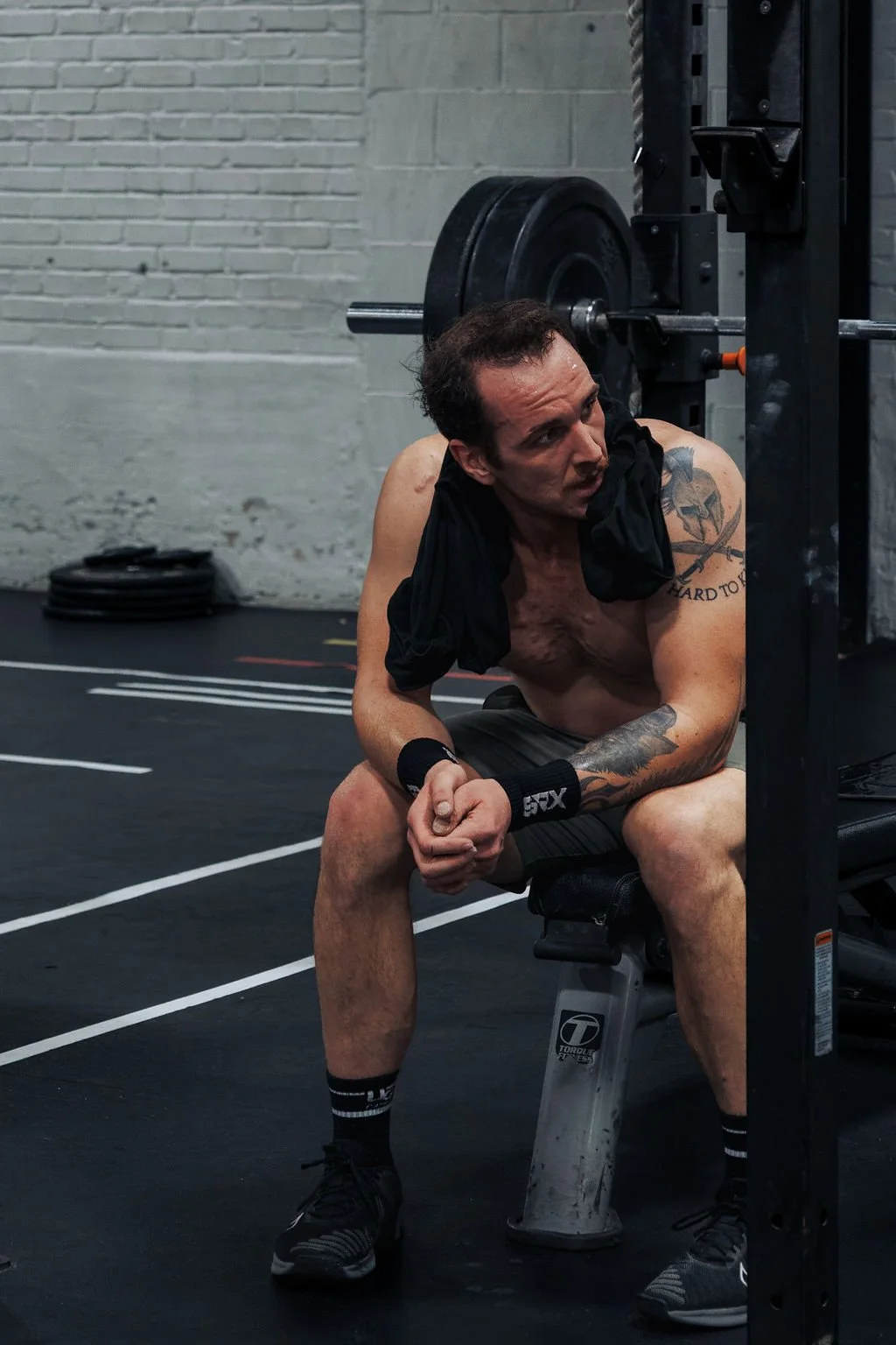 A shirtless man with tattoos sitting on a bench in a gym, resting after exercise, with weights and gym equipment in the background.