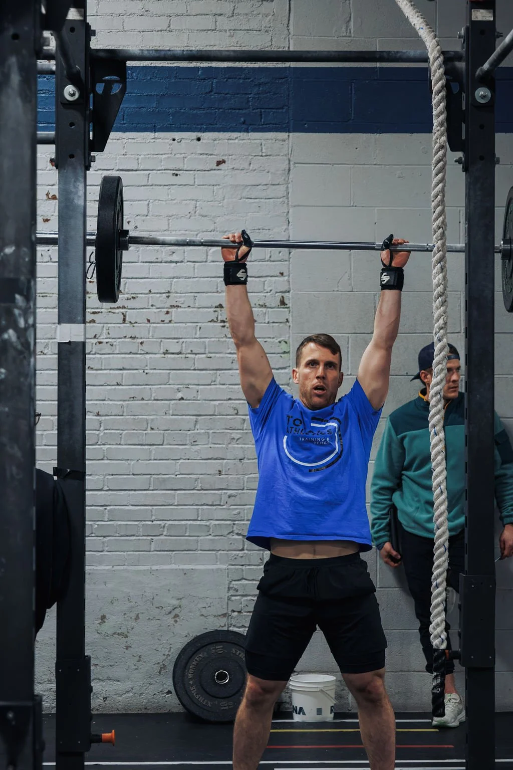 A man in a blue t-shirt and black shorts lifting a barbell overhead during a workout, standing in front of a brick wall in a gym.