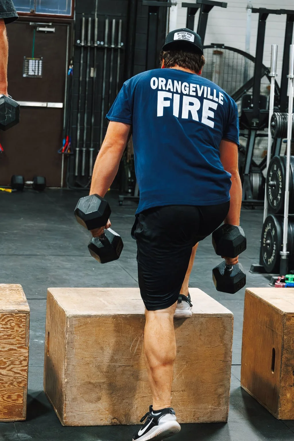 A person wearing a blue shirt with 'Orangeville Fire' on the back, black shorts, and a cap, lifting dumbbells while stepping onto a wooden box in a gym.