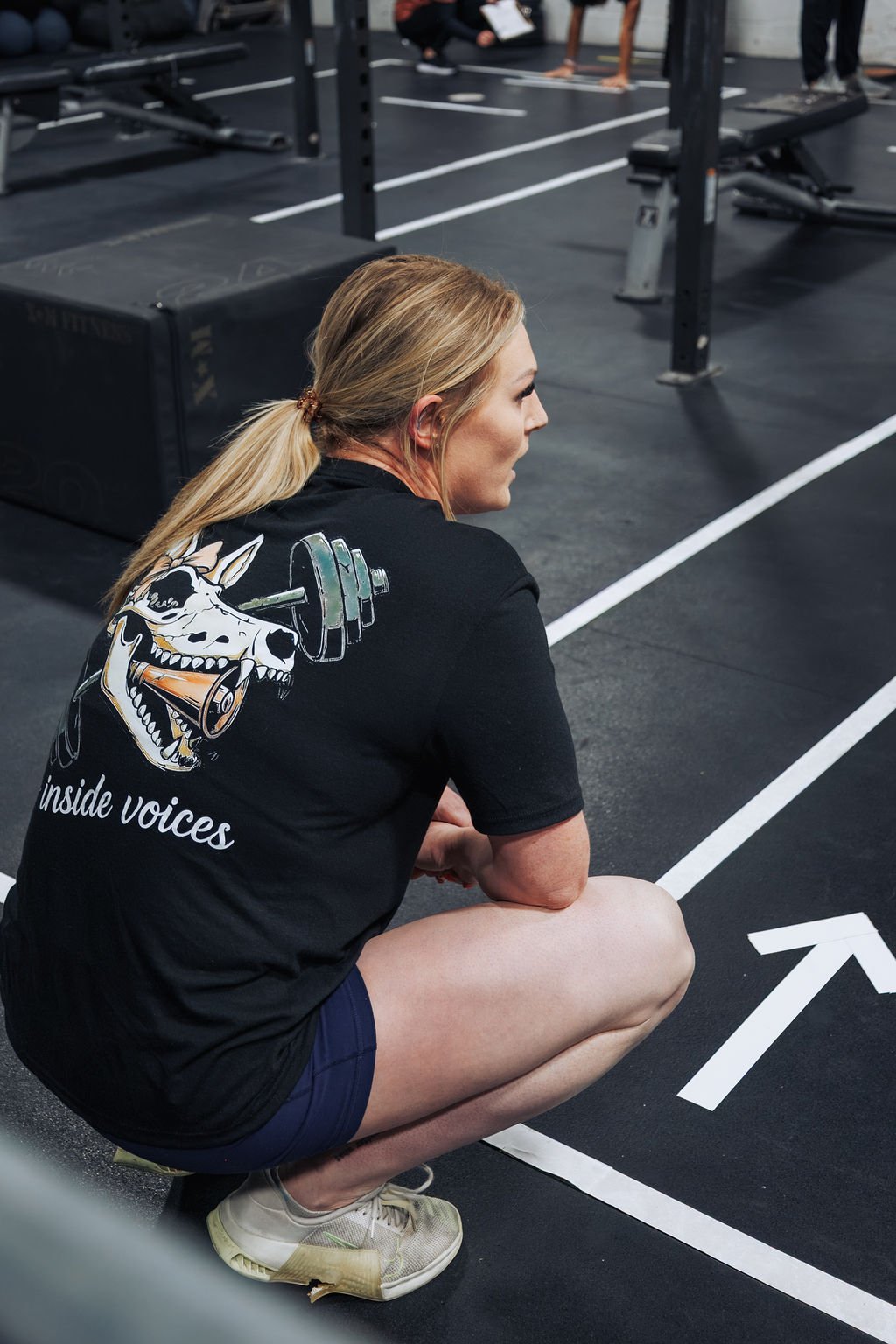 A woman with blonde hair tied back, sitting on the floor of a gym, wearing a black T-shirt with a graphic of a dog skull lifting a barbell and the text "inside voices." She is squatting with her arms resting on her knees, looking to the side.