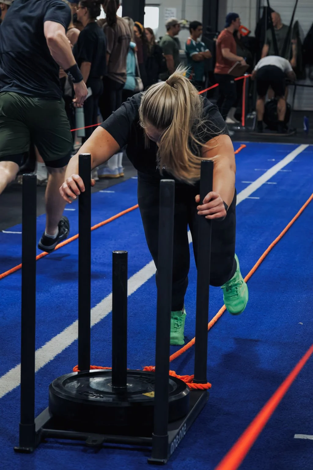 Woman in black workout attire pushing a sled with weights on a blue gym floor, with people in line and trainers in the background.