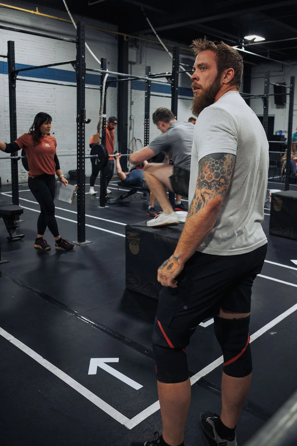 A man with a beard and tattoos on his arm is holding a black box inside a gym. In the background, a woman is observing, and several people are doing weightlifting exercises, including a person on a rowing machine and another doing squats with a barbe