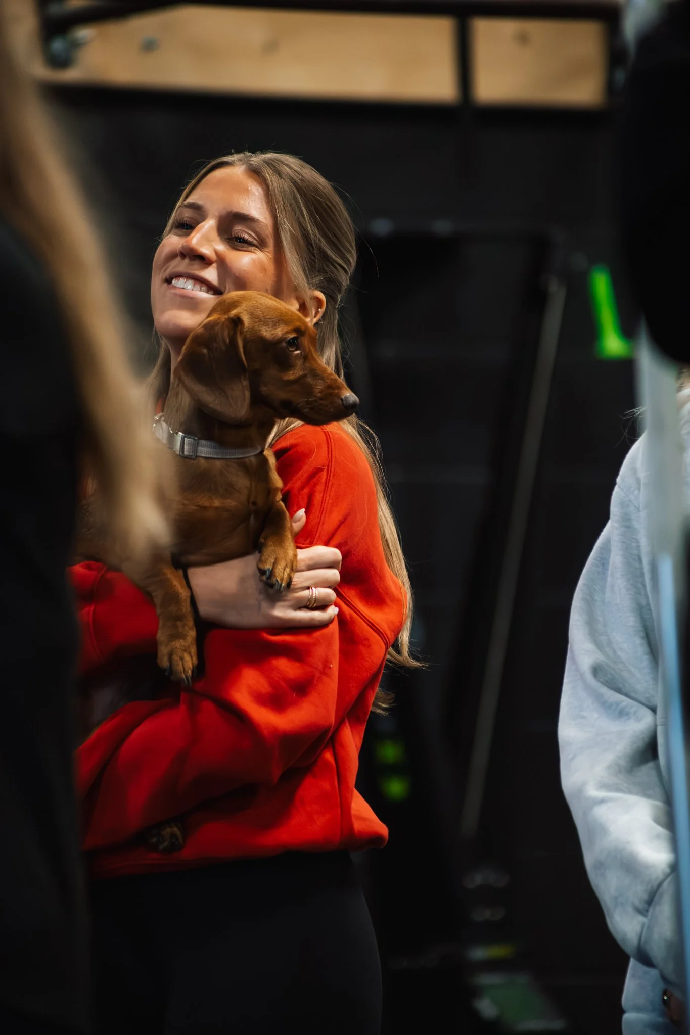 A woman with long blonde hair wearing a red hoodie is holding a brown dachshund puppy close to her chest. She is smiling and appears to be in a social setting with other people around.
