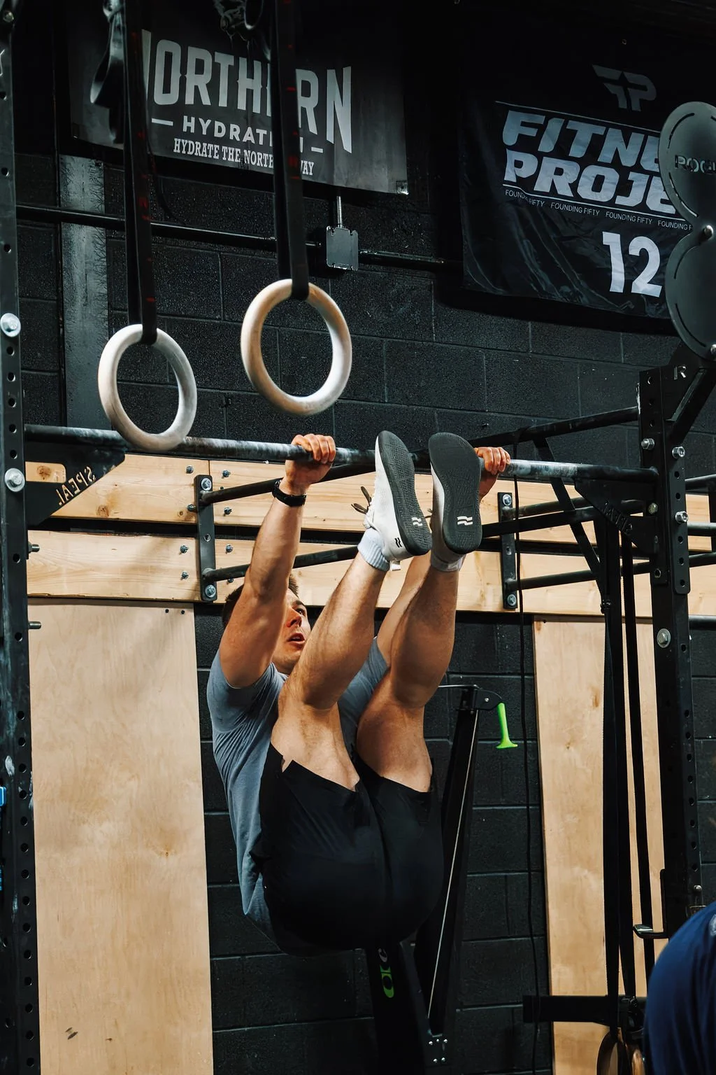 A man performing a bar muscle-up at the gym, holding onto the bar with his legs raised and feet pointed, with gym gymnastic rings and fitness banners visible in the background.