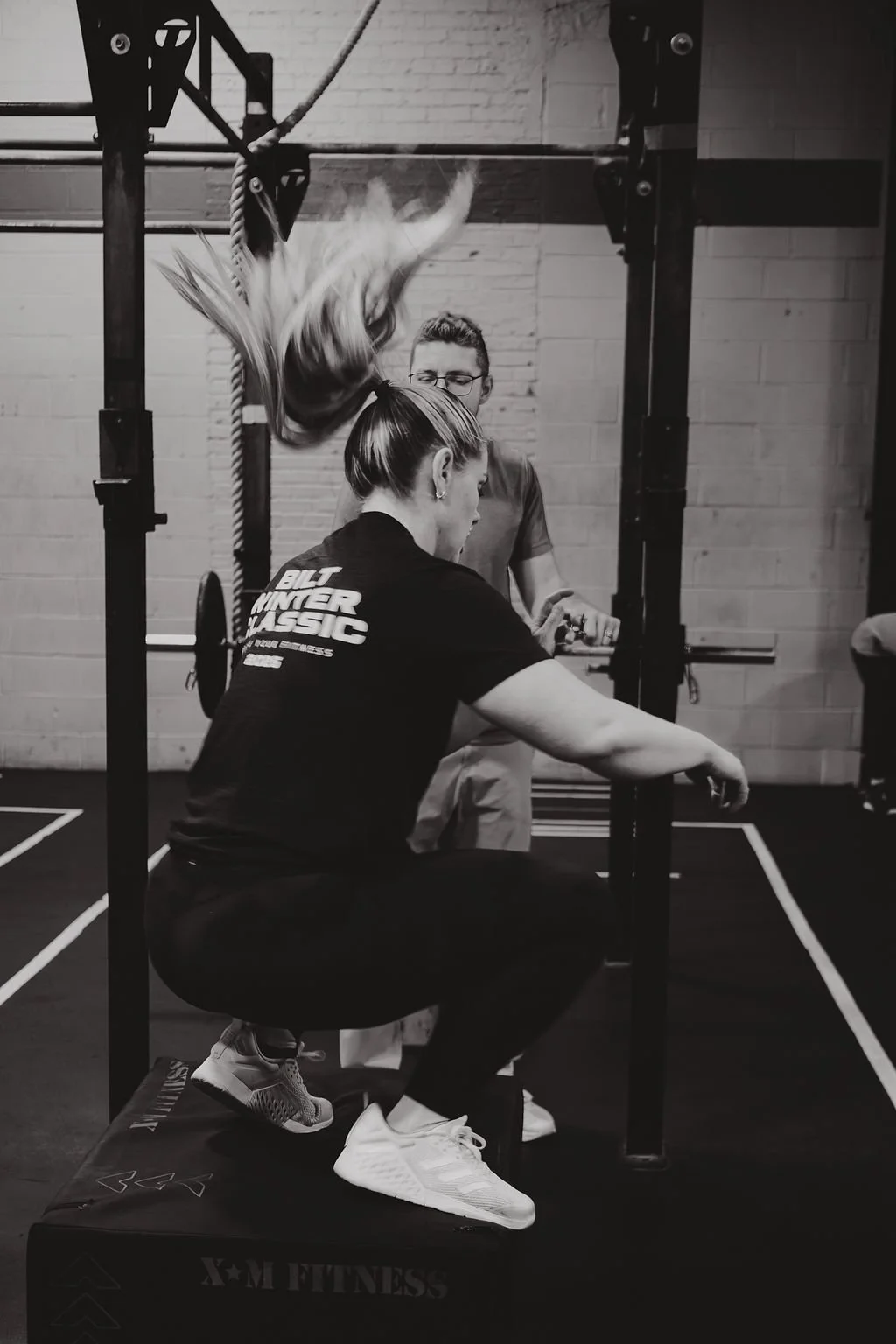 A woman with long hair in a ponytail is squatting on a platform, preparing to lift weights at a gym with a trainer standing behind her. The trainer appears to be assisting or guiding her during the workout.