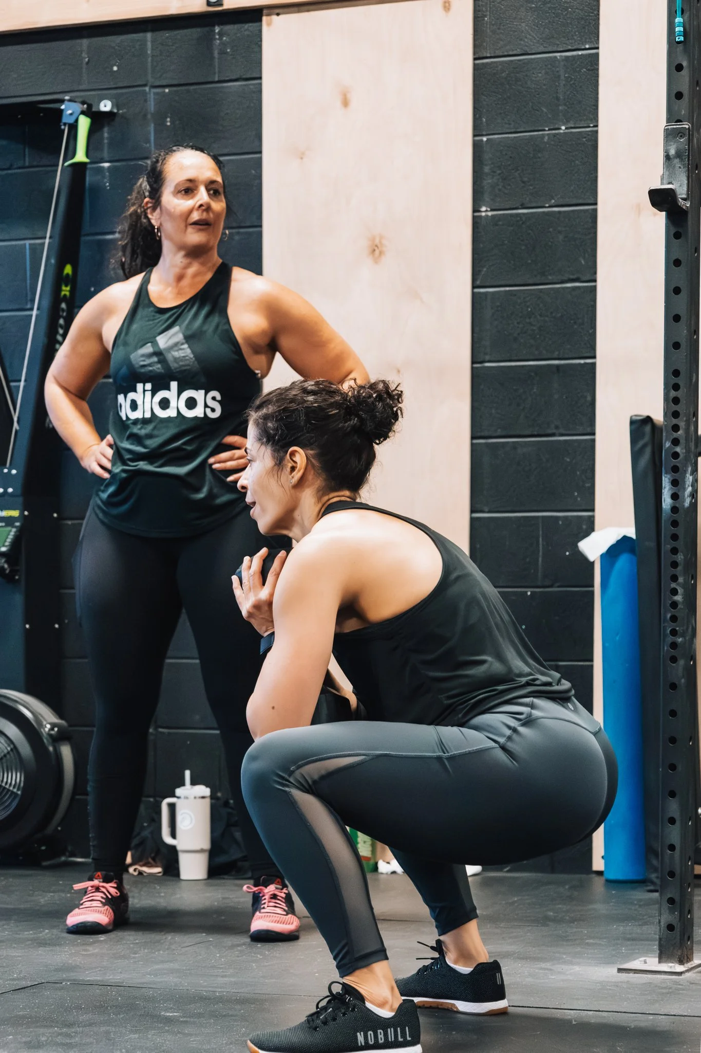 Two women in athletic clothing having a conversation in a gym. One is standing with hands on hips, and the other is crouching.
