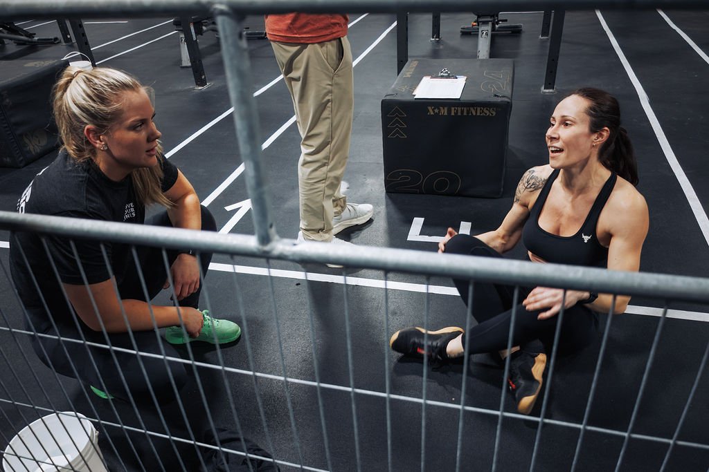 Two women athletes are sitting on the gym floor behind a metal fence, having a conversation. One is squatting and looking at the other, who is sitting with her legs crossed, smiling. A bench and a plyometric box are in the background.